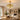 Luna ceiling fan light fixture with a brushed gold metal frame and white curved blades, installed in a bright, contemporary living room with a wooden dining table in the foreground.